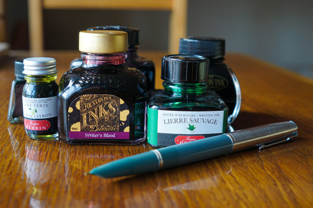 Several bottles of red and green coloured ink on a polished wooden desk top with a vintage Parker 51 fountain pen.
